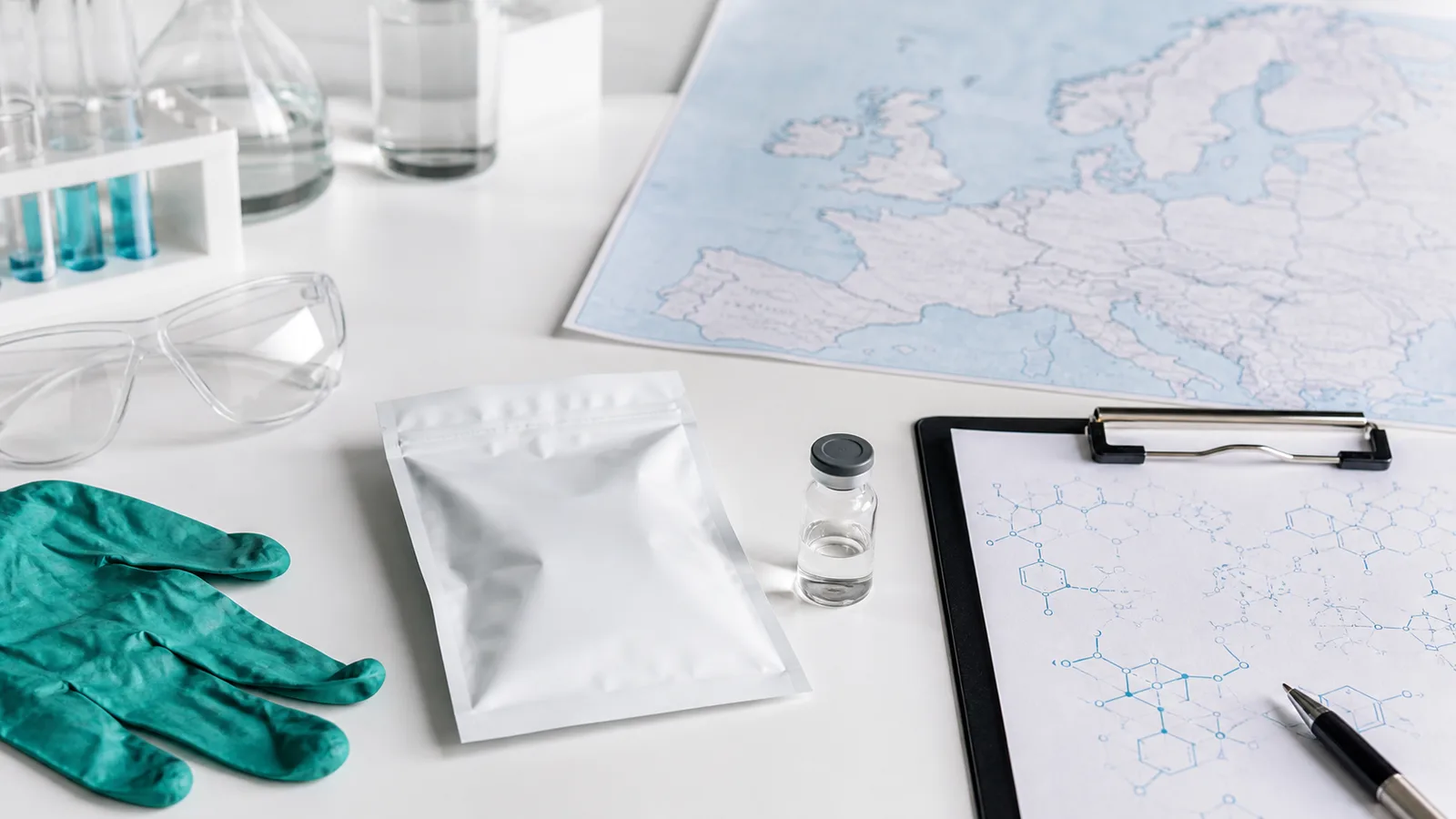 Suljettu tutkimusnäytepussi, laboratoriovälineet ja Euroopan kartta puhtaalla työpinnalla.